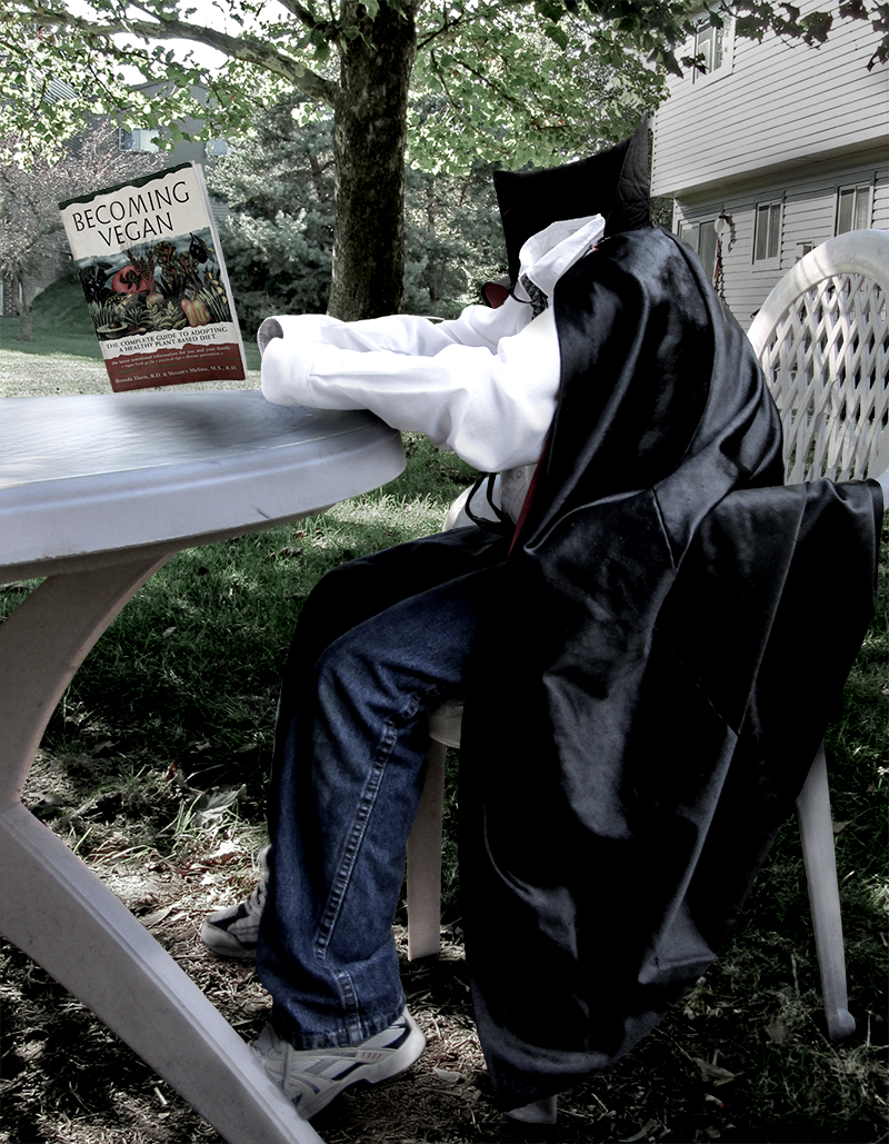Invisible boy in a vampire cape, sitting at a table outside, reading a book about the vegan diet.